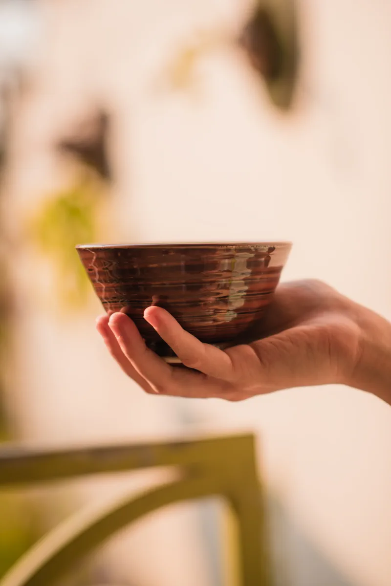 A hand supports a dark red ceramic bowl from below, showing its layered reflective glaze and foot