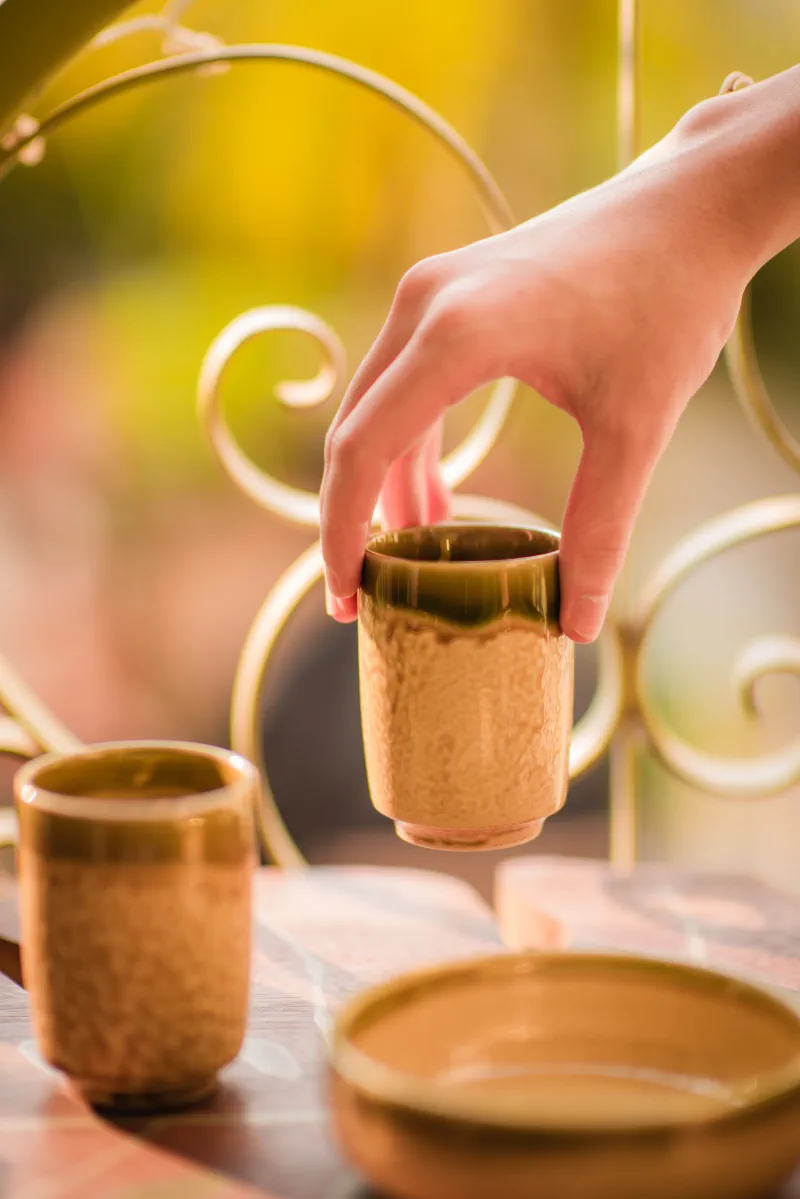 A hand lifts a tall speckled ceramic cup with an olive-glazed rim above the rest of the set
