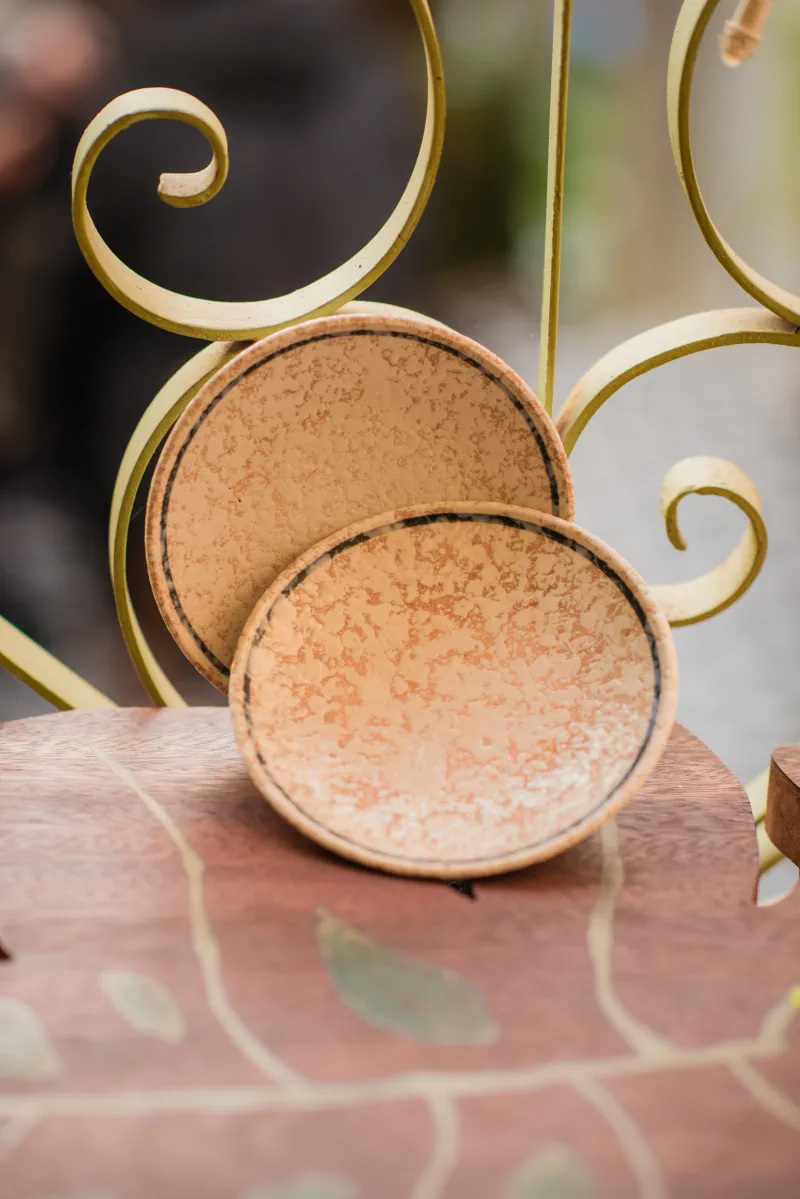 Two cream speckled ceramic plates lean upright among pale wrought-iron curves in sunlight