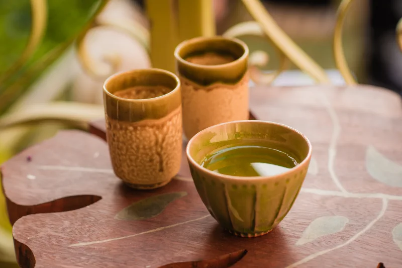 A green-glazed tea bowl filled with water sits before two tall textured cups on a leaf-shaped wooden table