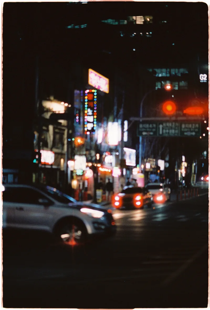 Cars pass through a soft field of neon signs and red taillight halation at night