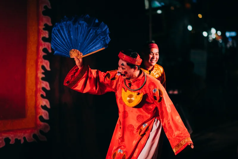 A comic tuồng performer in a red costume snaps open a blue fan while another performer laughs behind him