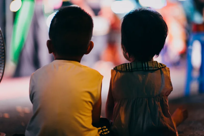 Two children seen from behind sit close together and watch a brightly lit tuồng stage on a Hà Nội street