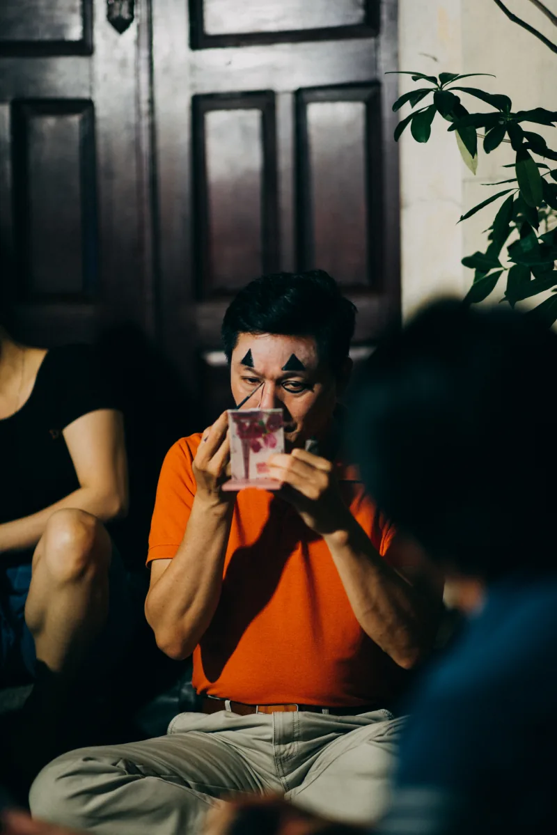 A performer in an orange shirt applies stage makeup while looking into a small hand mirror in a dark doorway