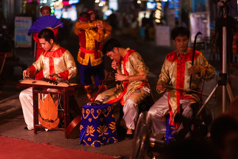 Three musicians in cream and red costumes play traditional instruments beside a tuồng stage on the street