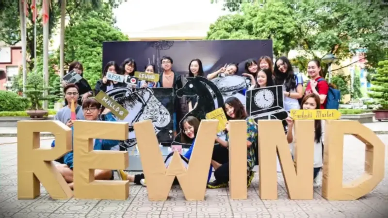 The production crew crowds behind giant cardboard REWIND letters, holding hand-drawn props in a campus yard