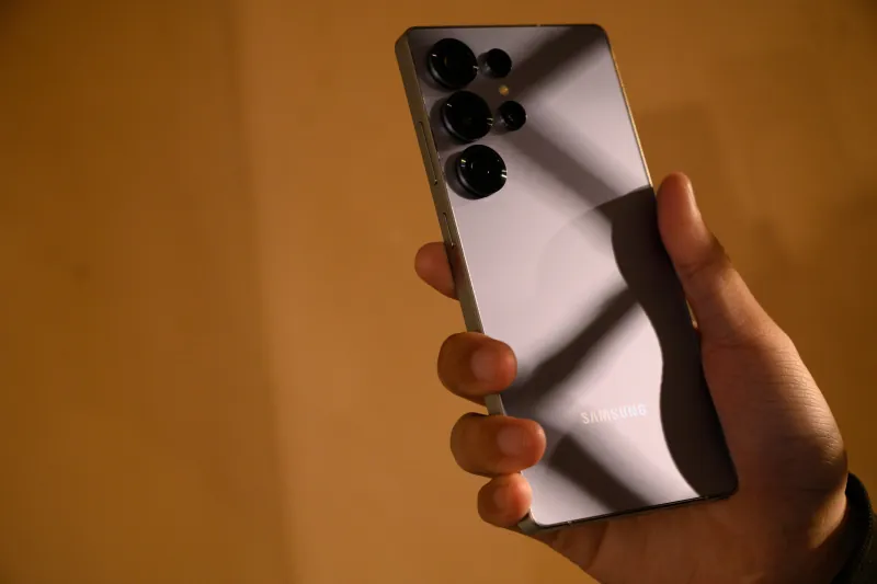 A silver Galaxy S25 Ultra held in warm light, with window shadows crossing its back and camera rings