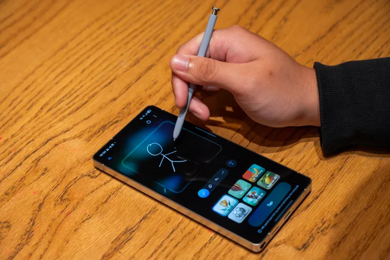 A hand sketches a figure with the S Pen on a Galaxy S25 Ultra lying on a wooden table