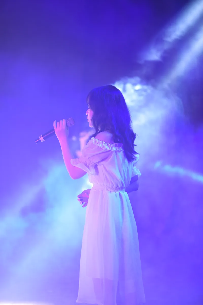 A singer in a white dress stands in profile inside blue-violet stage haze