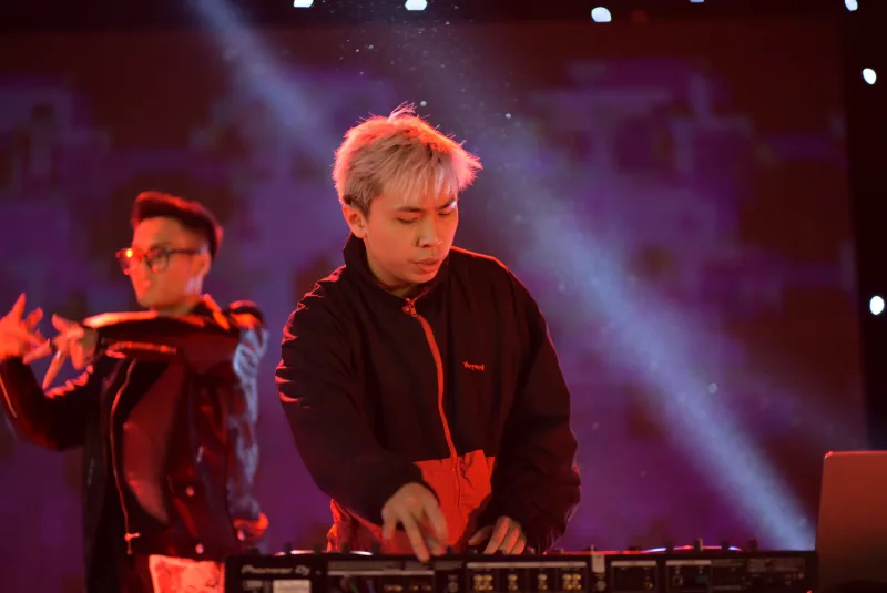 A DJ leans over a controller under red light as another performer moves behind him