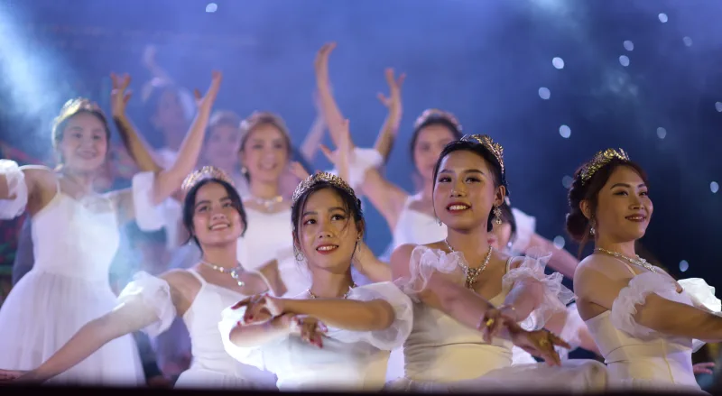 A line of dancers in white performs under blue haze and pin lights