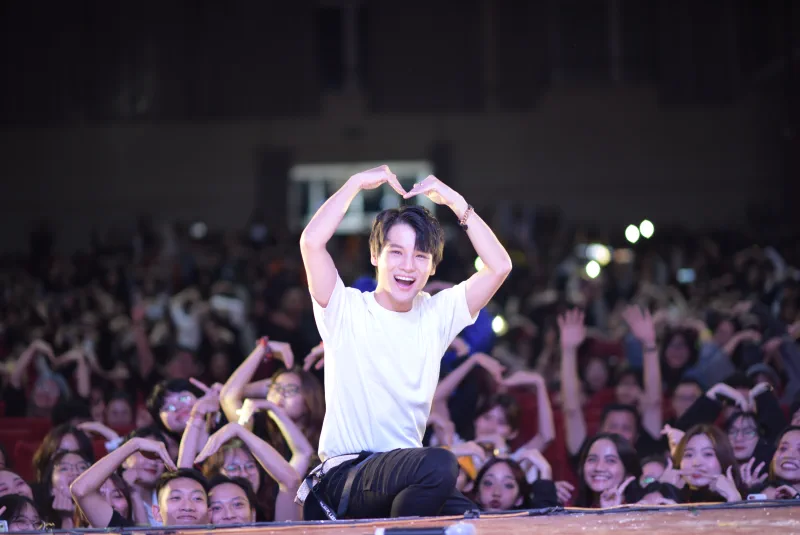 A smiling performer forms a heart above his head while the front rows echo the gesture