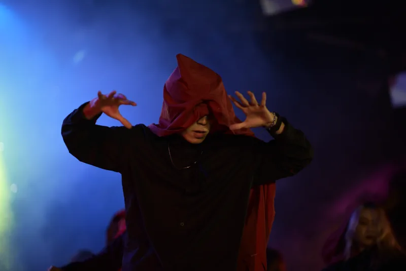A hooded performer raises both hands while a red veil falls across their face